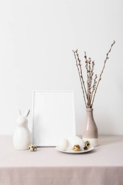 Blank photo frame, plate with Easter eggs, rabbit and pussy willow branches on table near light wall