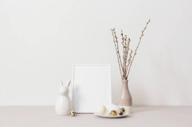 Blank photo frame, plate with Easter eggs, rabbit and pussy willow branches on table near light wall