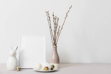 Blank photo frame, plate with Easter eggs, rabbit and pussy willow branches on table near light wall