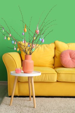 Masadaki vazoda Paskalya yumurtalarının olduğu ağaç dalları ve odanın renk duvarının yanındaki kanepe.