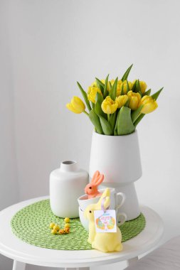 Laleli vazolar, fincanlar ve aydınlık duvarın yanındaki masada tebrik kartı olan Paskalya tavşanları.