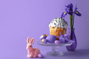 Şirin Paskalya tavşanı, pasta, boyalı yumurta ve mor arka planda çiçekli vazo.