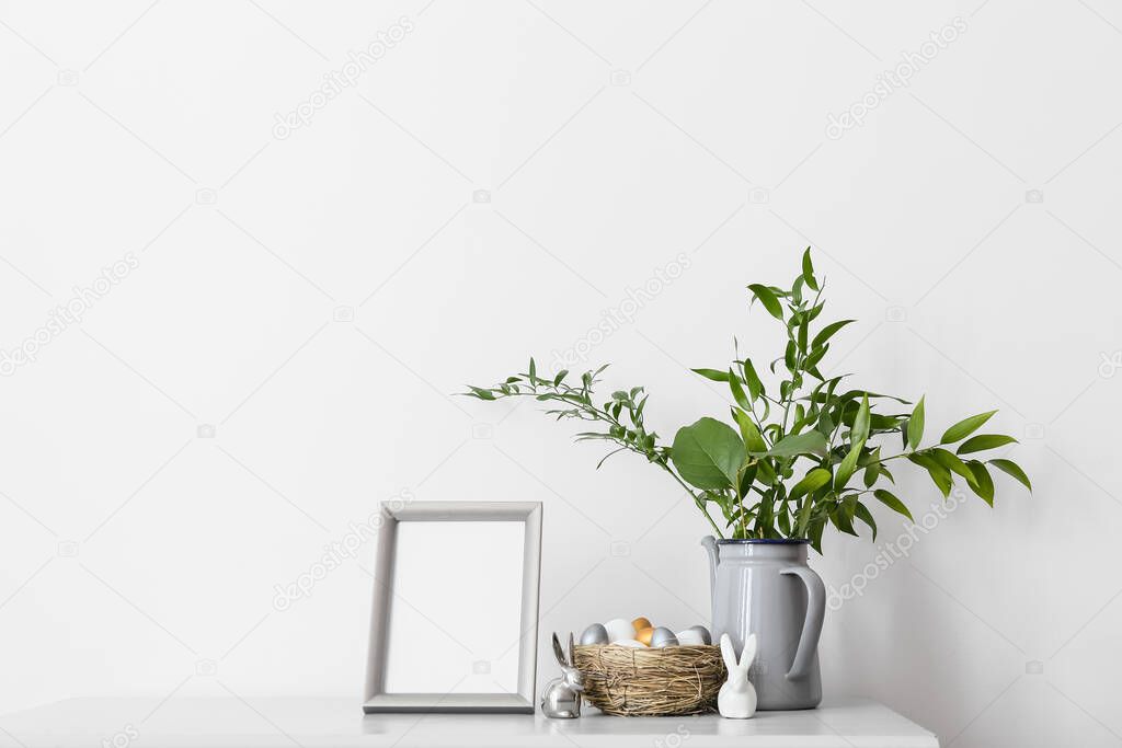 Blank photo frame, nest with Easter eggs and different plant branches on table near light wall