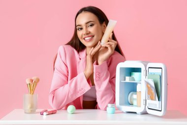 Küçük buzdolabı ve kozmetik ürünleriyle güzel genç bir kadın renk arka planında masada oturuyor.