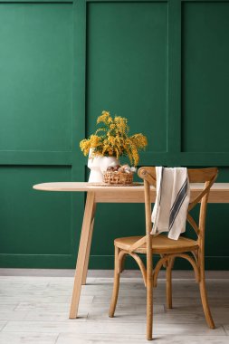 Mimoza çiçekli vazo, sepet ve yeşil duvarın yanındaki yemek masasında paskalya yumurtası.