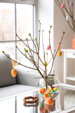 Oturma odasındaki cam masanın üzerinde ağaç dalları ve Paskalya yumurtaları olan vazo.
