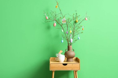 Vazoda Paskalya yumurtalarıyla süslenmiş ağaç dalları ve renkli duvarın yanındaki ahşap masada fincanlar.