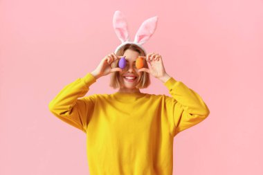 Tavşan kulaklı ve pembe arka planda Paskalya yumurtası olan komik genç bir kadın.