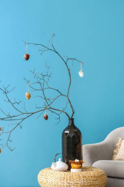 Paskalya kutlaması için süslenmiş ağaç dalları olan vazo mavi duvarın yanındaki masada lezzetli çörekler ve çaydanlık.