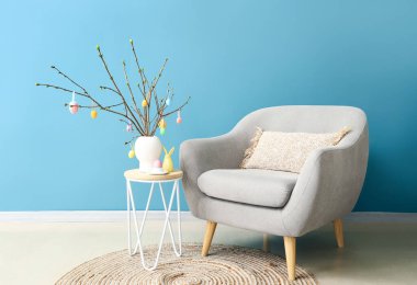 Masadaki vazoda Paskalya yumurtaları ve mavi duvarın yanındaki koltukla süslenmiş ağaç dalları.