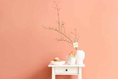 Lezzetli çörekler, fincanlar, ağaç dalları ve renkli duvarın yanındaki masada vazoda Mutlu Paskalyalar yazan tebrik kartı.