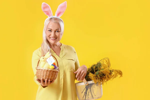 Ältere Frau Mit Hasenohren Osterkorb Und Blumen Auf Gelbem Hintergrund — Stockfoto