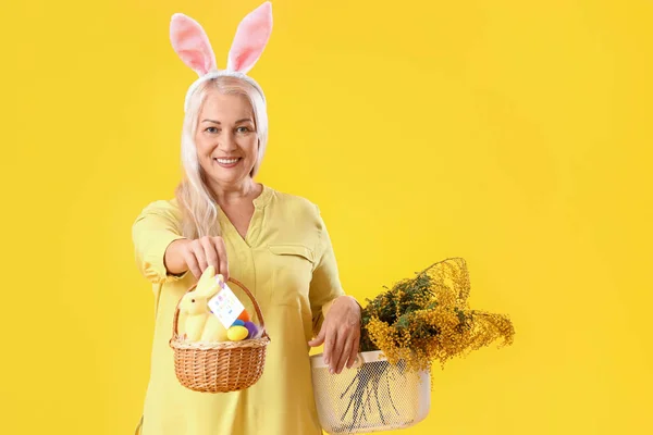 Ältere Frau Mit Hasenohren Osterkorb Und Blumen Auf Gelbem Hintergrund — Stockfoto
