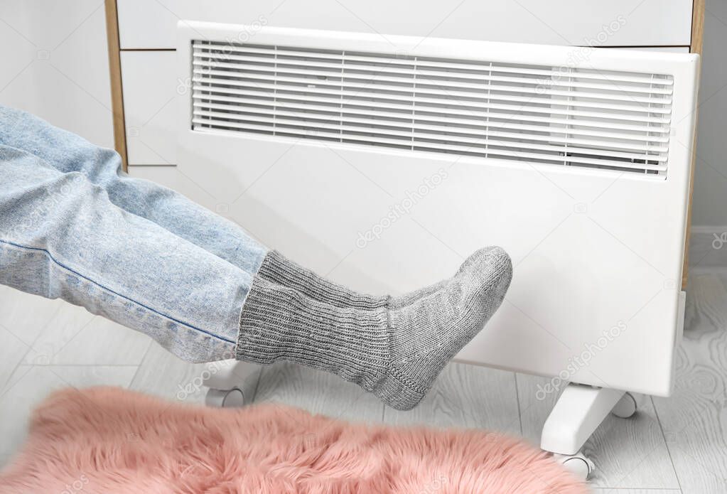 Woman in warm socks near radiator at home. Concept of heating season