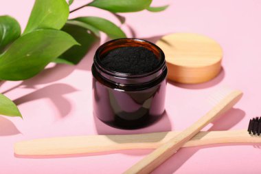 Kavanozda aktif kömür diş tozu ve pembe arka planda diş fırçası var.