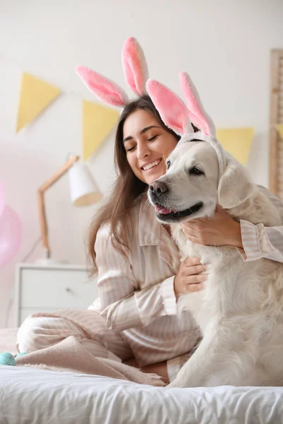 若い女性と自宅でベッドの上にウサギの耳を持つ愛らしい犬 イースターのお祝い — ストック写真