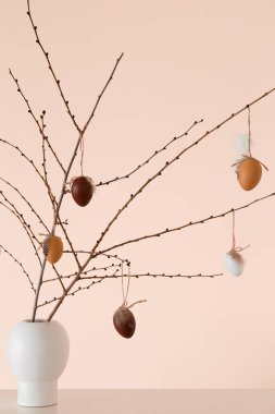 Ağaç dalları ve bej duvarın yanındaki rafta Paskalya yumurtaları olan vazo.