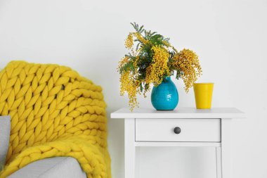 Mimoza çiçekli vazo ve bardakla ışık duvarının yanındaki masada.