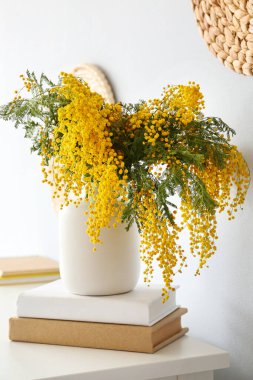 Mimoza çiçekli vazo ve ışık duvarının yanındaki masada kitaplar.