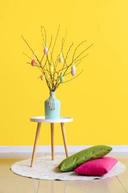 Masanın üzerinde paskalya yumurtaları ve sarı duvarın yanında yastıklar olan ağaç dalları.