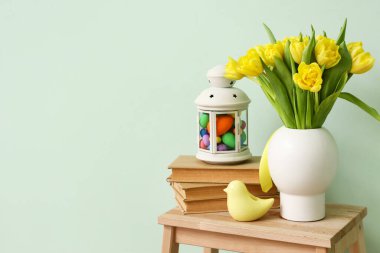 Laleli vazo, kitap yığını, Paskalya yumurtalı fener ve renkli duvarın yanındaki merdivende duran kuş.