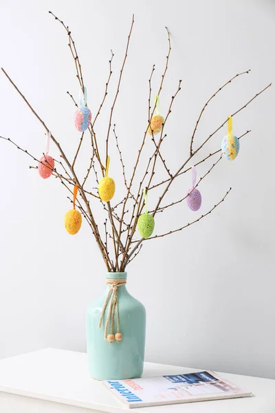 Vase with tree branches, colorful Easter eggs and magazine on shelf near light wall