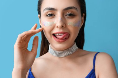 Yaratıcı makyajlı kadın yüzüne dokunuyor ve mavi arka planda dilini gösteriyor.