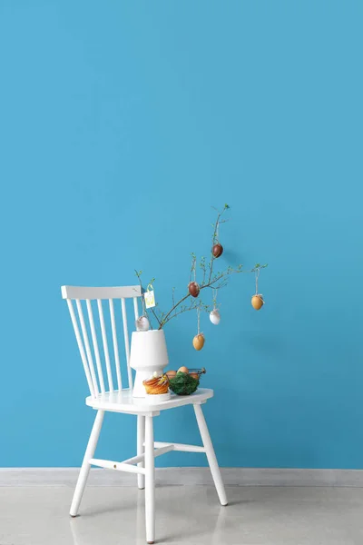 部屋のカラーウォールの近くの椅子にイースターエッグの木の枝 おいしいパンとバスケットと花瓶 — ストック写真