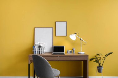 Laptop, çerçeve ve sarı duvarın yanında parlayan lambayla modern işyeri
