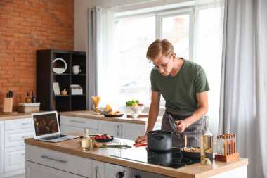 Genç adam mutfakta video dersi takip ederken çorba yapıyor.