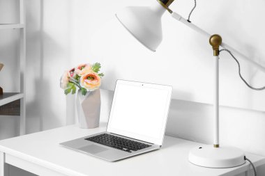 Dizüstü bilgisayarlı modern işyeri, vazoda çiçekler ve ışık duvarının yanında lamba.