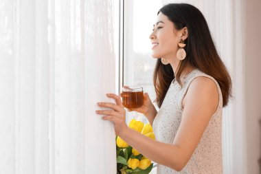 Güzel genç Asyalı kadın pencerenin yanında sıcak çay içiyor.