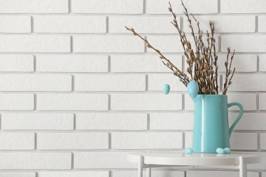 Jug with pussy willow branches and painted Easter eggs on table near white brick wall