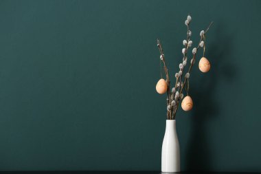 Vase with pussy willow branches and painted Easter eggs on table near green wall