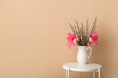 Jug with pussy willow branches and tulip flowers on table near beige wall