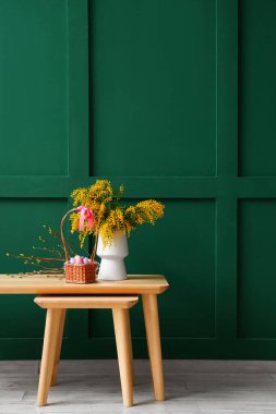 Mimoza çiçekli vazo, ağaç dalları, yeşil duvarın yanındaki sepetin içinde paskalya yumurtası.