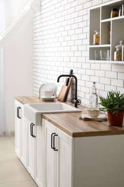 Lavabolu tezgahlar, ev bitkisi ve mutfak aletleri beyaz tuğla duvarın yanında.