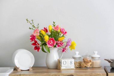 Güzel çiçekli sürahi ve 8 Mart tarihli küp takvimi mutfak tezgahında, ışık duvarının yanında. Uluslararası Kadınlar Günü kutlamaları