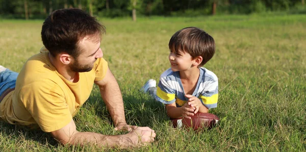 小男孩拿着橄榄球和他的父亲躺在公园的绿草上 — 图库照片