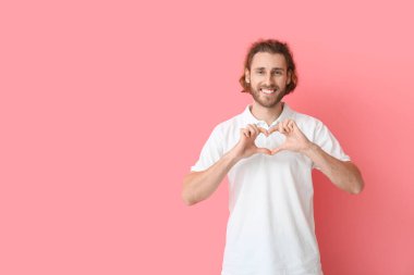 Yakışıklı, polo tişörtlü, elleri pembe arka planda kalp yapan genç bir adam.