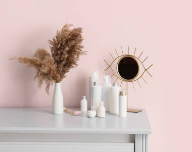 Farklı kozmetik ürünleri, yüz masajı aracı, ayna ve pampa otları, renkli duvarın yanındaki çekmecelerde.