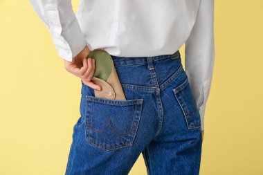 Kadın, sarı arka planda kot pantolon cebine tekrar kullanılabilir adet pedi koyuyor.