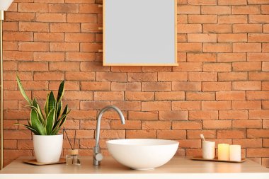 Lavabolu masa, ev bitkisi, kamış ayırıcı, mum ve diş fırçası tuğla duvarın yanında.