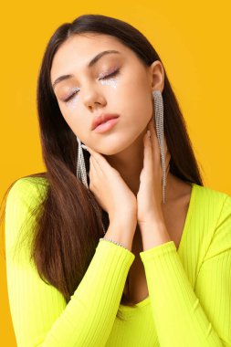 Yaratıcı makyajlı ve gözleri kapalı bir kadın sarı arka planda boyuna dokunuyor.