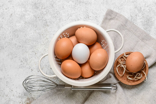 Colander with fresh chicken eggs and whisk on light background