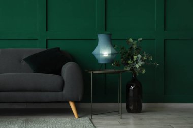 Kahve masasında parlayan lamba, okaliptüslü vazo ve yeşil duvarın yanındaki kanepe.