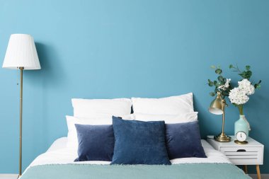 Rahat bir yatak, komodinin üstünde çiçekli vazo ve mavi duvarın yanında modern lamba.
