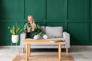 Yeşil duvarın yanında oturmuş çay içen olgun bir kadın.