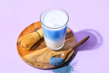 Bir bardak mavi kibrit çayı, mor arka planda toz ve kaşık.