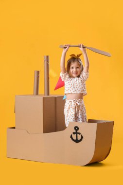 Sarı arka planda karton gemiyle oynayan sevimli küçük kız.